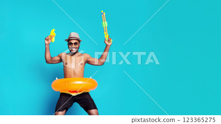 Cheerful young black man jumping with two water guns over blue background, wearing swimming clothes and inflatable ring 123365275