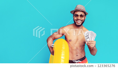 A shirtless African American man in a straw hat and sunglasses smiles broadly while holding a stack of US dollar bills and a bright yellow inflatable ring against a light blue background. A shirtless African American man in a straw hat and sunglasses smiles broadly while holding a stack of US dollar bills and a bright yellow inflatable ring against a light blue background. 123365276