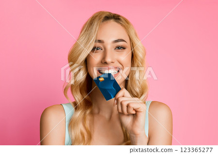 A smiling woman with long blonde hair is holding a credit card in front of her mouth. She is wearing a light blue tank top and is looking directly at the camera. The background is a solid pink color. A smiling woman with long blonde hair is holding a credit card in front of her mouth. She is wearing a light blue tank top and is looking directly at the camera. The background is a solid pink color. 123365277
