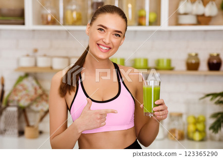 A woman in a pink sports bra holds a glass of green smoothie in a kitchen. 123365290