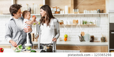 A family is cooking together in a modern kitchen. The father is holding their young child, who is reaching for a piece of fruit. The mother is stirring a pot on the stove, copy space 123365298