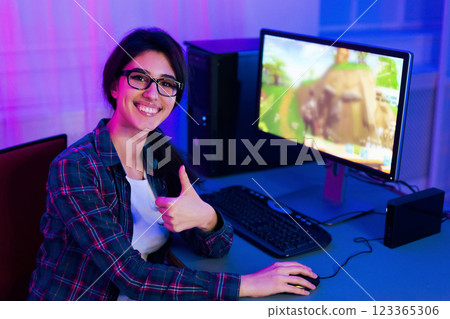 A woman wearing glasses smiles while playing a video game on a computer. Her thumbs up gesture shows her enjoyment. She sits at a desk lit with purple and blue lighting in what appears to be a home 123365306
