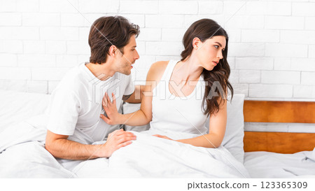 A young man and woman are sitting in bed, arguing. The woman is looking away, while the man is reaching towards her, seemingly trying to make amends. 123365309