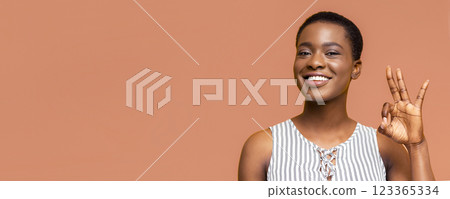 A young black woman with short black hair smiles broadly as she makes an okay sign with her right hand. The woman is wearing a sleeveless striped shirt. The background is a solid peach color. A young black woman with short black hair smiles broadly as she makes an okay sign with her right hand. The woman is wearing a sleeveless striped shirt. The background is a solid peach color. 123365334