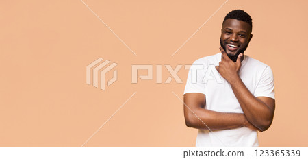 A young African American man in a white t-shirt smiles brightly while standing against a peach colored background. He has a thoughtful expression and his right hand is resting on his chin, copy space A young African American man in a white t-shirt smiles brightly while standing against a peach colored background. He has a thoughtful expression and his right hand is resting on his chin, copy space 123365339