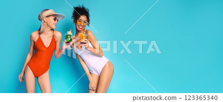 Two young women in swimsuits, one wearing a white swimsuit and the other a red one, are holding drinks in their hands and smiling at each other. The women are on a blue background, copy space Two young women in swimsuits, one wearing a white swimsuit and the other a red one, are holding drinks in their hands and smiling at each other. The women are on a blue background, copy space 123365340