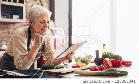 A senior woman in an apron uses a tablet to find a recipe in her kitchen. The countertop is filled with fresh produce, including cucumbers, tomatoes, bell peppers, and broccoli. 123365341