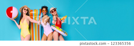 Positive young multiethnic girlfriends wearing swimsuits holding beach accessories, posing together on blue studio background, panorama with copy space Positive young multiethnic girlfriends wearing swimsuits holding beach accessories, posing together on blue studio background, panorama with copy space 123365356