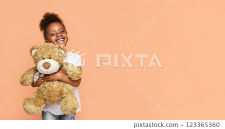 A little black girl with dark hair is smiling brightly as she holds a large, brown teddy bear in front of a peach-colored background. The girl is wearing a white t-shirt and blue jeans, copy space 123365360