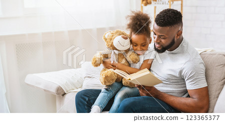 African American father and daughter are sitting on a couch in a living room, reading a book together. It is a heartwarming scene of a father and daughter bonding over reading. African American father and daughter are sitting on a couch in a living room, reading a book together. It is a heartwarming scene of a father and daughter bonding over reading. 123365377