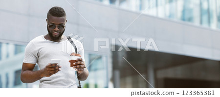 A man in sunglasses checks his phone while holding a cup of coffee, walking on a city street. 123365381