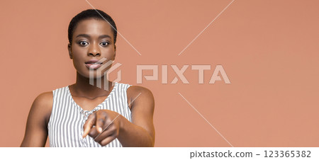 A young African American woman with short hair, wearing a striped tank top, stands in front of a light orange background and stares directly at the camera. She points a finger directly at the viewer 123365382
