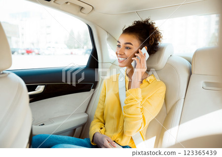 African American young woman wearing a yellow shirt sits in the back seat of a car and talks on her phone while looking out the window. The car appears to be parked in a city setting. 123365439