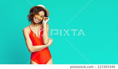 African American woman in a red one-piece swimsuit is smiling at the camera. She is wearing yellow sunglasses and a white hat. She is standing in front of a turquoise background, copy space 123365440