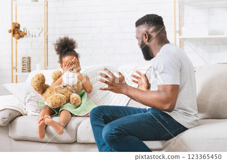 An African American father sits on a couch, smiling and playfully covering his eyes with his hands. His young daughter sits beside him, covering her eyes with her hands as well, holding a teddy bear 123365450