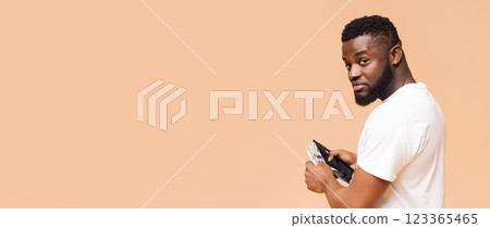 A young African American man wearing a white t-shirt stands against a beige background. He holds a wallet in his right hand, showing a glimpse of cash inside. He looks over his shoulder 123365465