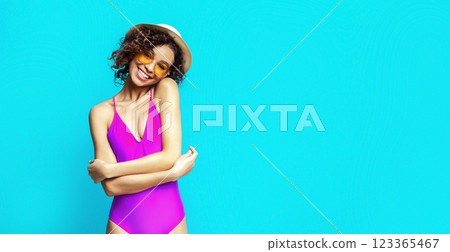 This image features African American smiling woman with curly brown hair wearing a pink swimsuit and a white hat. She is posing in front of a bright blue background, copy space 123365467