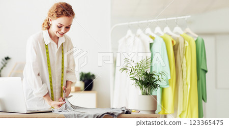 A young woman in a white shirt cuts fabric at a table in a home studio. She is wearing a measuring tape and is smiling. A young woman in a white shirt cuts fabric at a table in a home studio. She is wearing a measuring tape and is smiling. 123365475