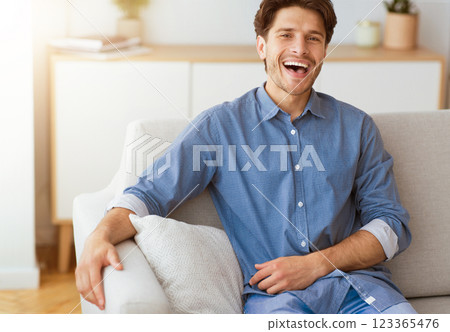 A man in a blue button-down shirt sits on a white couch and laughs. He is relaxed and looks happy. 123365476
