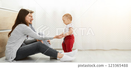 A young family of three are spending time together at home. The parents are sitting on the floor, smiling at their baby who is taking their first steps, copy space 123365488
