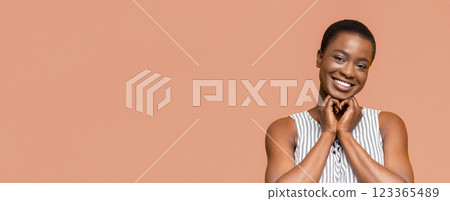 African American young woman with short hair is smiling and looking directly at the camera. She is making a heart shape with her hands in front of her chest 123365489