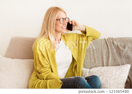 A woman in a yellow cardigan sits on a couch talking on her cell phone. She is smiling and looking at the phone. 123365490