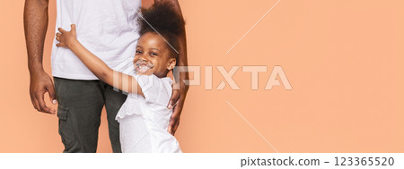 A little African American girl with curly hair is hugging her father from behind. She is smiling and looking directly at the camera. Her father is standing to the right of the frame, copy space A little African American girl with curly hair is hugging her father from behind. She is smiling and looking directly at the camera. Her father is standing to the right of the frame, copy space 123365520