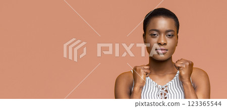 This image is a portrait of black woman with short, dark hair against a peach background. She is looking directly at the camera with a serious expression on her face and her fists are clenched. 123365544