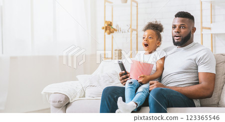 African American father and daughter are seated on a couch in a living room watching television. The young girl is holding a container of popcorn and looking intently at the television, copy space 123365546