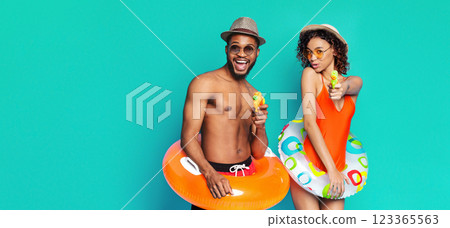 A happy couple is posing against a bright teal background. The man is wearing a brown fedora and sunglasses and holding a yellow water gun. He is holding an orange inflatable ring, copy space A happy couple is posing against a bright teal background. The man is wearing a brown fedora and sunglasses and holding a yellow water gun. He is holding an orange inflatable ring, copy space 123365563