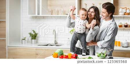 A happy family is playing in their modern kitchen while preparing food. The mother is holding up their young child, who is smiling and reaching out. Father is standing next to them, looking lovingly A happy family is playing in their modern kitchen while preparing food. The mother is holding up their young child, who is smiling and reaching out. Father is standing next to them, looking lovingly 123365568