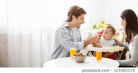 A smiling father feeds his baby while his wife watches nearby. They are enjoying a breakfast together at home, with a glass of orange juice and a bowl of snacks on the table, copy space 123365578