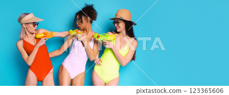 Three young women are posing in swimsuits on a blue background. They are holding water guns and looking playful. They appear to be having fun at a summer pool party or beach. Three young women are posing in swimsuits on a blue background. They are holding water guns and looking playful. They appear to be having fun at a summer pool party or beach. 123365606