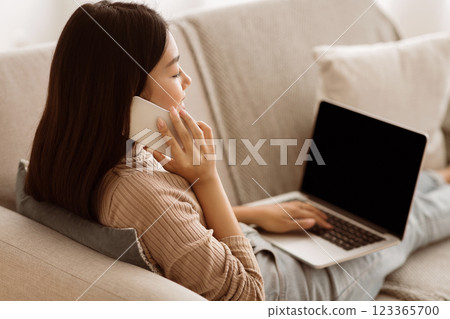 A woman is sitting on a couch, talking on her phone while using a laptop. Her hand is resting on the laptop keyboard. 123365700