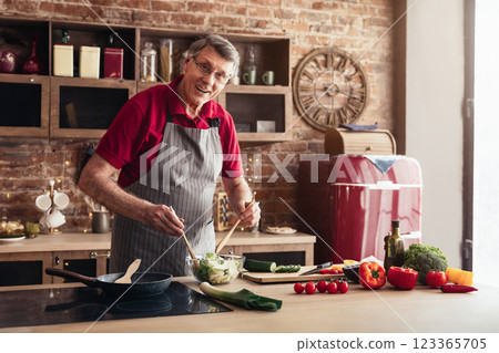A senior man wearing a grey apron and a red shirt is preparing a salad in his modern kitchen. He is standing at the counter, holding a wooden spoon in each hand, and stirring a bowl of green salad A senior man wearing a grey apron and a red shirt is preparing a salad in his modern kitchen. He is standing at the counter, holding a wooden spoon in each hand, and stirring a bowl of green salad 123365705
