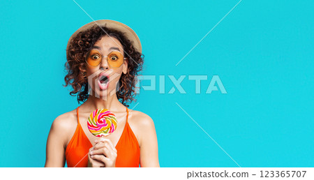 Young black woman in an orange swimsuit holding a colorful lollipop and looking surprised, with a blue background behind her. Her mouth is open, and she is wearing sunglasses and a straw hat. 123365707