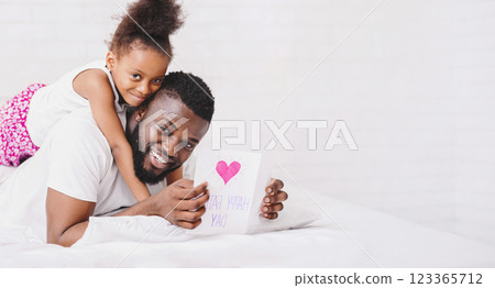 Black father is lying in bed holding a card that has a pink heart and handwritten words on it, smiling at the camera. His daughter is sitting on his back, her arms wrapped around his neck 123365712