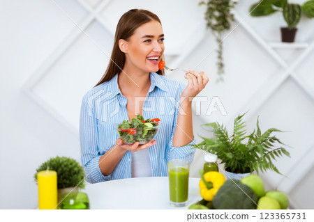 A woman smiles as she eats a healthy salad with a fork, enjoying a fresh green juice in a white interior setting. A woman smiles as she eats a healthy salad with a fork, enjoying a fresh green juice in a white interior setting. 123365731