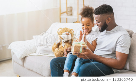African American young girl, sitting on her fathers lap, opens a gift. Happy family celebrating birthday together at home, copy space 123365740