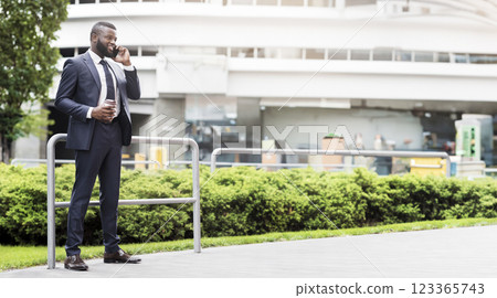Handsome african american businessman using mobile phone during coffee break outdoors, standing against the railing. Full length Handsome african american businessman using mobile phone during coffee break outdoors, standing against the railing. Full length 123365743