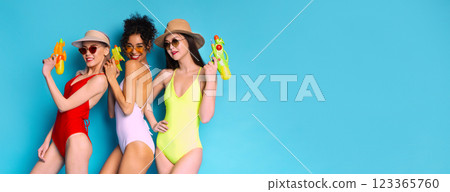 Three women wearing swimsuits and hats are holding water guns while standing against a bright blue background. They are smiling and appear to be enjoying themselves. 123365760