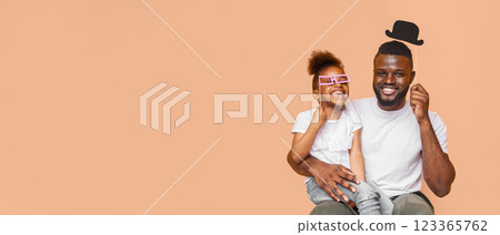 Happy black father and daughter posing with photo booth props. The father is holding his daughter on his lap. The father has a black top hat on his head and the daughter is wearing pink glasses. 123365762