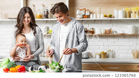 A young family is preparing a salad in a modern kitchen. The mother is holding their baby, who is playfully biting a red pepper. The father is stirring a bowl of salad with wooden spoons A young family is preparing a salad in a modern kitchen. The mother is holding their baby, who is playfully biting a red pepper. The father is stirring a bowl of salad with wooden spoons 123365768