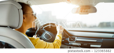 African American woman in sunglasses smiles while driving a car, with her hand on the steering wheel. The view outside the car window is blurred, with a bright sunny sky in the background. 123365882