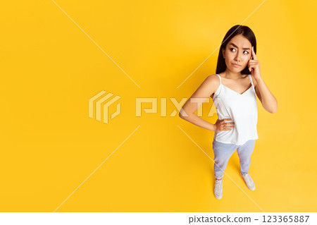 Asian young woman standing in front of a yellow background. She is wearing a white tank top and blue jeans. She is looking to the side with a thoughtful expression on her face, top view, copy space 123365887
