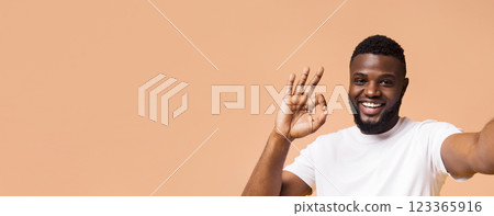 A smiling Black man with a beard and a white t-shirt is making the okay sign with his hand, looking directly at the camera. He is standing against a light brown background, copy space A smiling Black man with a beard and a white t-shirt is making the okay sign with his hand, looking directly at the camera. He is standing against a light brown background, copy space 123365916