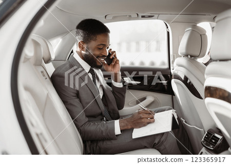 African American businessman in a suit sits in the backseat of a luxury car and makes notes on a notepad while speaking on his phone. He looks focused and professional. 123365917