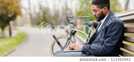 Black businessman uses his laptop in a park, seated on a bench with his bicycle beside him. The mood is serene and efficient, showcasing outdoor work-life balance, panorama Black businessman uses his laptop in a park, seated on a bench with his bicycle beside him. The mood is serene and efficient, showcasing outdoor work-life balance, panorama 123365941