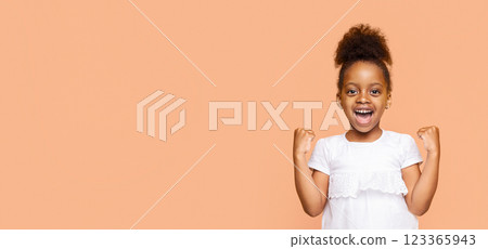 A little Black girl with a big smile and her fists raised in the air celebrates a victory or achievement. She is wearing a white shirt and her dark hair is styled in a high ponytail, copy space 123365943
