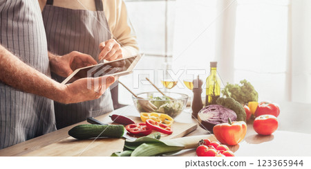 An elderly couple wearing aprons stand in their kitchen, looking at a tablet together. Kitchen is cozy and homey, with exposed brick walls, wooden shelves, and a vintage clock, cropped 123365944